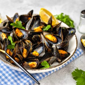 Mussels served in a white bowl with parsley and lemon wedges on a checkered cloth; oil and salt nearby.