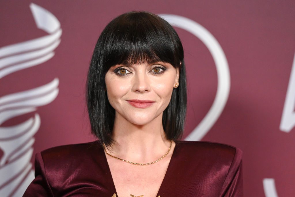 Person standing, smiling slightly, in maroon attire with a short necklace; set against a maroon backdrop featuring abstract white lines.
