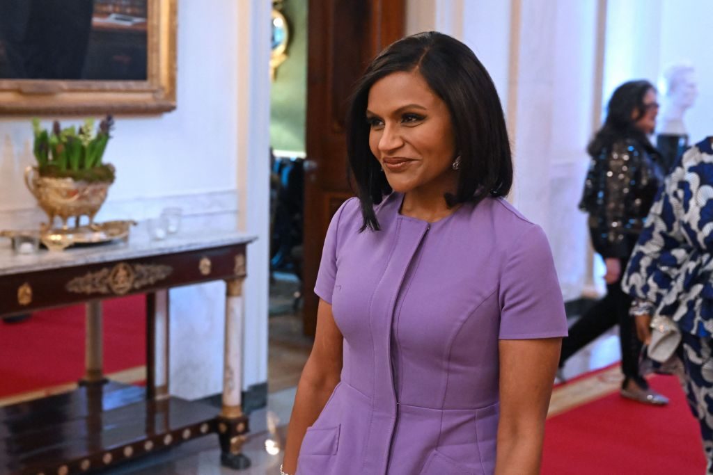 A person in a purple dress smiles while standing in an elegantly decorated room with red carpet and ornate furniture.