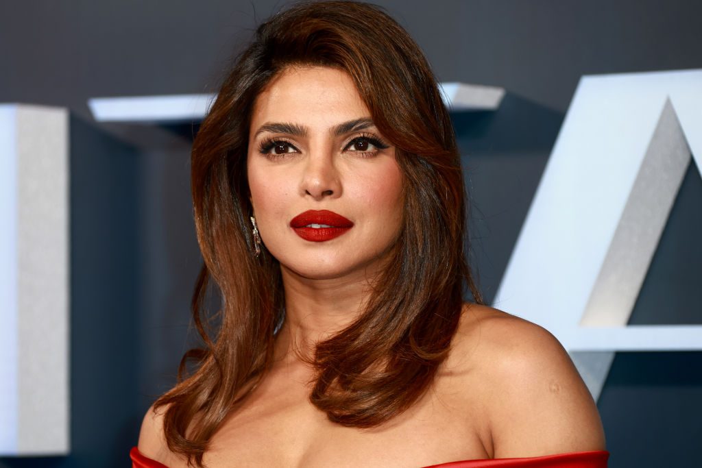 A woman with long hair and red lipstick stands against a dark backdrop at an event, wearing a red off-shoulder outfit.