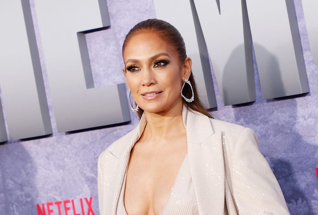 A woman stands smiling, wearing statement earrings and a light blazer, against a Netflix-themed backdrop displaying text.