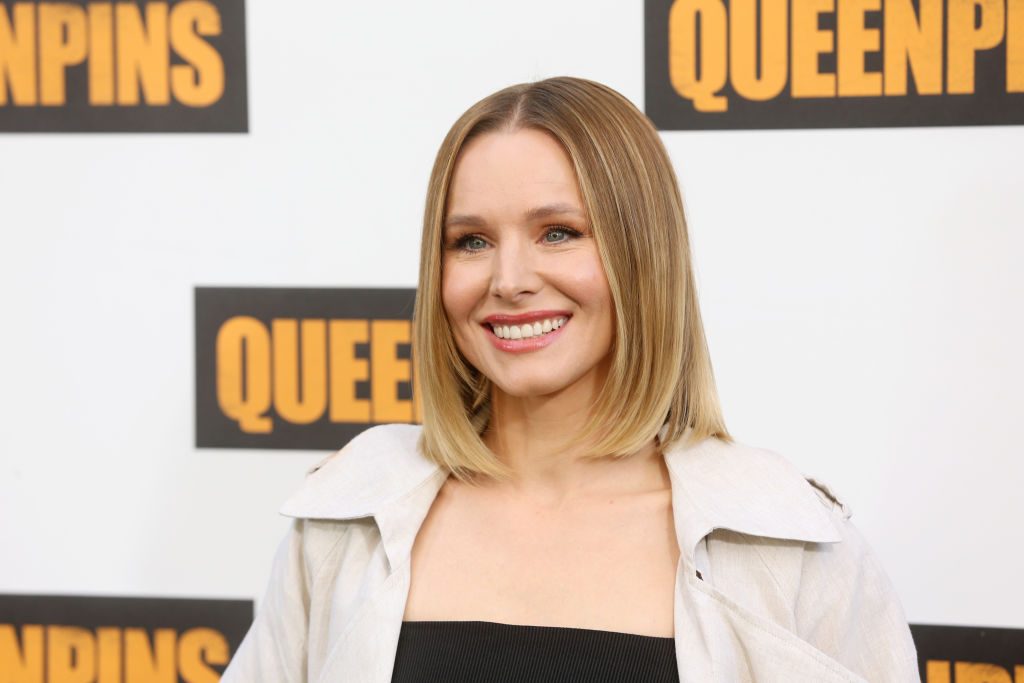 A person smiles, wearing a light-colored jacket, against a backdrop with the word "QUEENPINS" repeated in bold, yellow letters.
