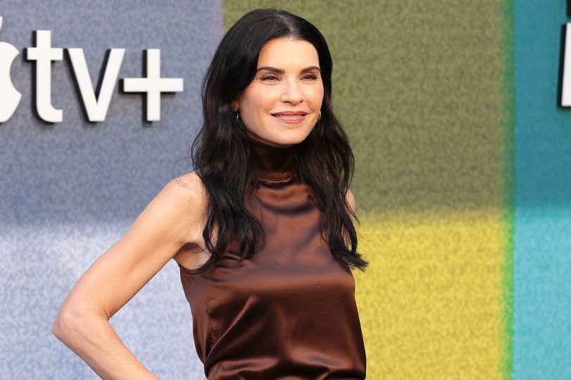 A person stands smiling, wearing a sleeveless brown top. The background features a colorful wall and "tv+" text on the left.