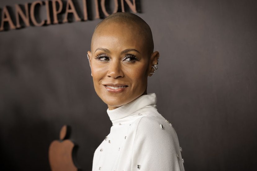 Person looking right, wearing a white high-neck shirt with embellishments, against a dark background featuring the word "EMANCIPATION" and an apple logo.