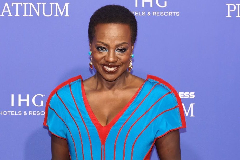 Smiling woman stands, wearing a bright blue and red dress. Purple backdrop displays text: "IHG", "HOTELS & RESORTS", "PLATINUM" partially visible.