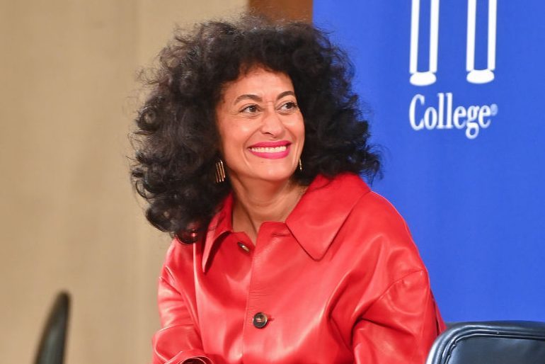 A person sits smiling in a red jacket, in front of a blue banner with partial text "College," indoors.