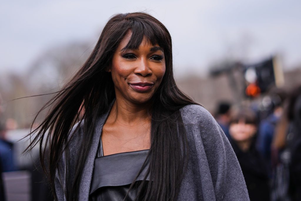 A person smiles while walking outside, with blurred crowd and trees in the background, wearing a dark outfit and long jacket.