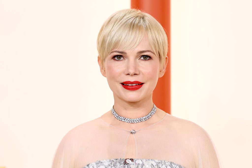 A person with short blond hair smiles, wearing a silver necklace and dress, standing against a soft, light background.