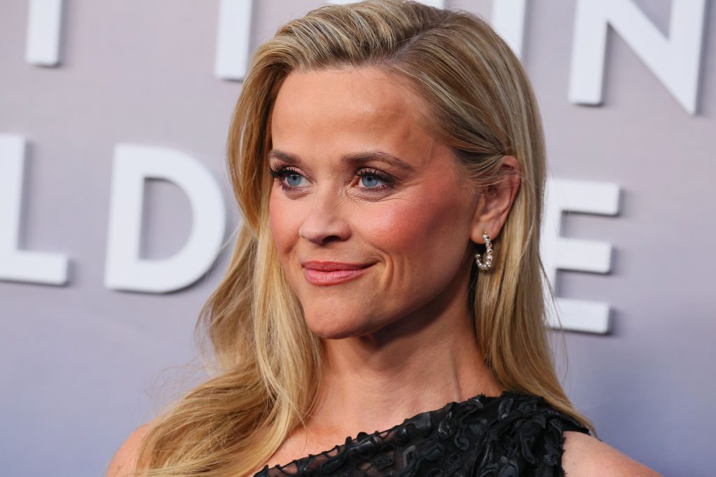 Person smiling slightly, looking to the side; wearing pearl earrings and a black textured dress. Background features large, partially visible white text.
