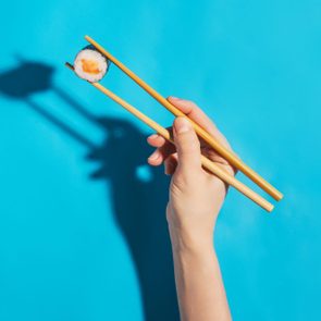 Chopsticks hold sushi against a bright blue background, casting a shadow of a dinosaur silhouette.