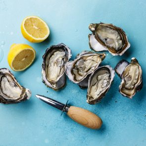 Oysters rest opened on a blue surface next to a lemon sliced in half and an oyster knife with a wooden handle.