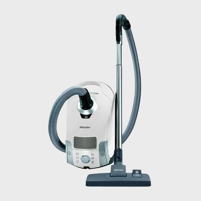 Vacuum cleaner stands upright; hose attached to base; gray context suggests a modern, sterile setting. The vacuum is labeled with 