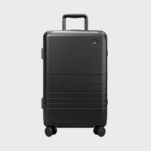 A black suitcase stands upright with handle extended, positioned against a plain gray background. Small logo reads 
