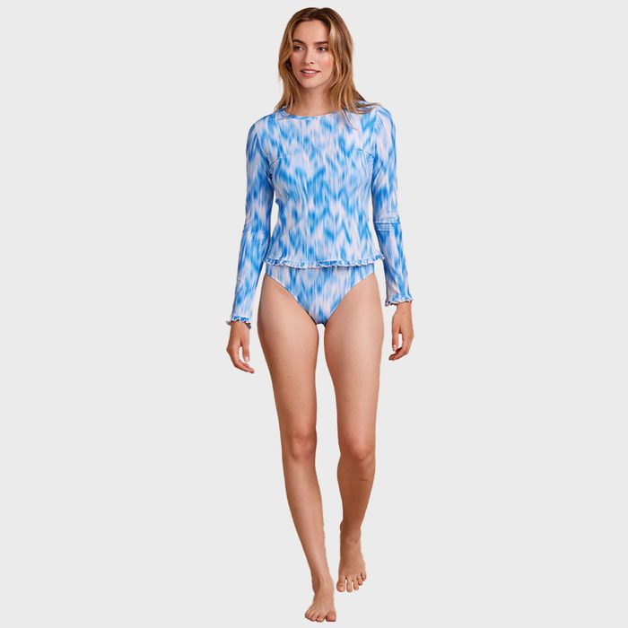 Woman models blue patterned long-sleeve swimsuit, standing, against plain background.