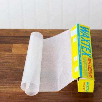 Wax paper roll partially unspooled from yellow box on wooden table, labeled "WAXTEX WAXED PAPER."