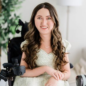 Person smiling in a floral dress sits in a wheelchair, surrounded by a plant and a lamp in a bright room.