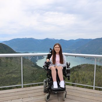 A person sits in a wheelchair, smiling, on a wooden deck overlooking a scenic mountainous landscape with a lake.