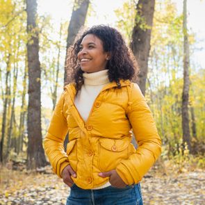 A person stands smiling, hands in pockets, wearing a yellow jacket, surrounded by sunlight-filtered autumn trees.