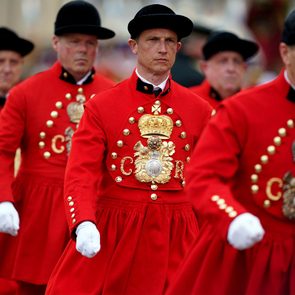 Men wear red uniforms with black hats and march in formation, adorned with gold buttons and insignias, in a formal setting.