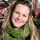 A smiling person wears a green scarf outdoors, with blurred market stalls and red flowers in the background.