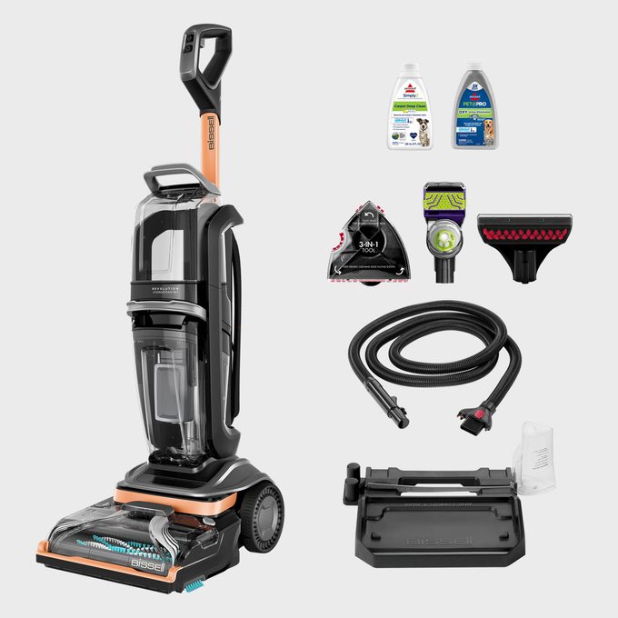 Vacuum cleaner stands upright, surrounded by various attachments and two bottles of cleaning solution against a plain background.