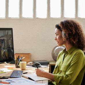 Woman with headphones participates in video call, writing in a notebook at a desk with a computer, phone, and colorful stationery.