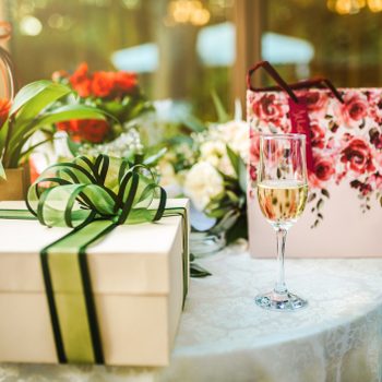 Gift box with green ribbon sits on table beside flowers and wine glass, set against a blurred outdoor garden background.