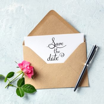 Card displays "Save the date," held by a brown envelope; a pink rose and pen lie beside it on a textured surface.