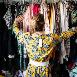 Person stands with raised arms, browsing clothes in a colorful, packed wardrobe. Tags hang from a yellow patterned dress worn by the person.