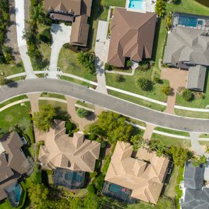 Road curves through suburban neighborhood; houses, driveways, and greenery seen from above in a sunny, landscaped environment.