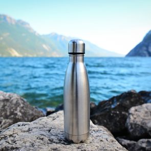 Metal water bottle stands on rocks near a lake, surrounded by distant mountains under a clear blue sky.