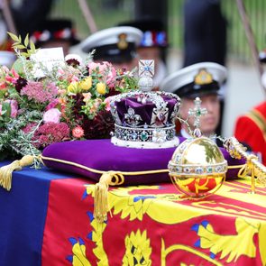 Crown and ceremonial items rest atop a draped casket adorned with vibrant flowers, surrounded by uniformed individuals in a formal setting.