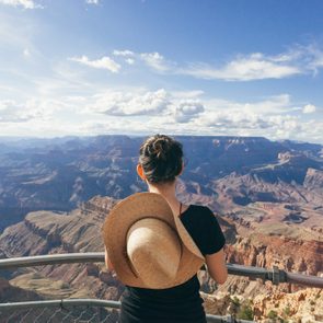 Person stands, holding a hat, overlooking a vast canyon landscape under a partly cloudy sky.