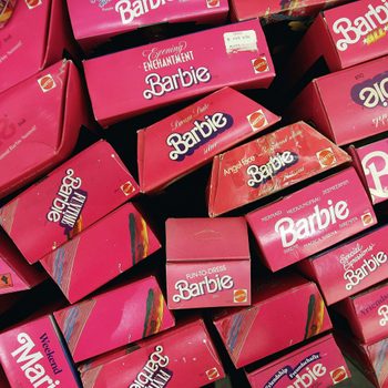 Barbie doll boxes, stacked haphazardly, feature bright pink packaging with various model names and the Mattel logo.