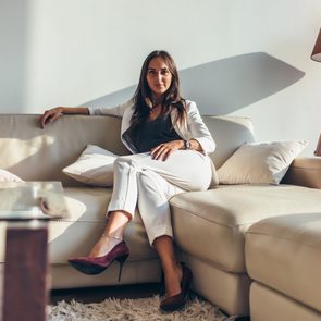 A woman sits relaxed on a cream-colored sofa, wearing a white suit, in a sunlit room with a floor lamp and glass table nearby.