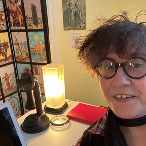 Person smiling, posing near vintage phone, lamp, open laptop on a desk; tarot cards and retro poster on wall, yellow room.
