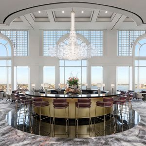 A large round bar is surrounded by stools, beneath an elegant chandelier, in a bright room with arched windows and a city view.
