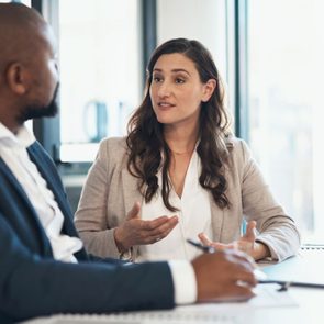 A woman gestures while talking to a man in a suit, seated inside an office with a laptop on the table.