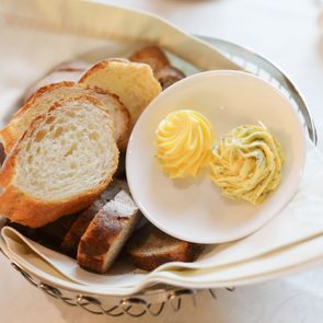 Sliced bread rests in a basket beside swirled butter portions on a small white plate, set on a tablecloth.