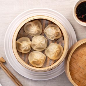 Steamed dumplings rest in a bamboo basket beside a small bowl of soy sauce, chopsticks, and a white spoon on a light wooden surface.