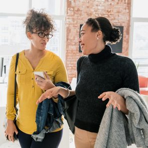 Two women walk while conversing, one holding a phone and jacket, in a bright office with desks and large windows.