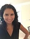 A person is smiling in a brightly lit room, wearing a black sleeveless top with curly hair, hand resting on hip.