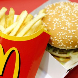 French fries in a red carton with a yellow logo beside a sesame seed burger on a red surface.