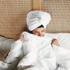 Person with towel on head, smiling under white duvet, resting against a woven wooden headboard in bed.