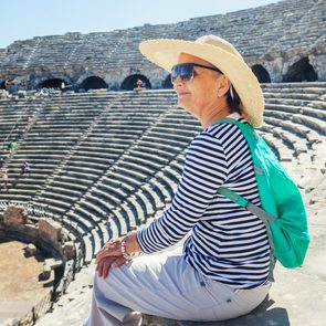 Woman in a sunhat sits observing ancient amphitheater steps, wearing sunglasses, striped shirt, and green backpack; sunlit stone seating surrounds her.