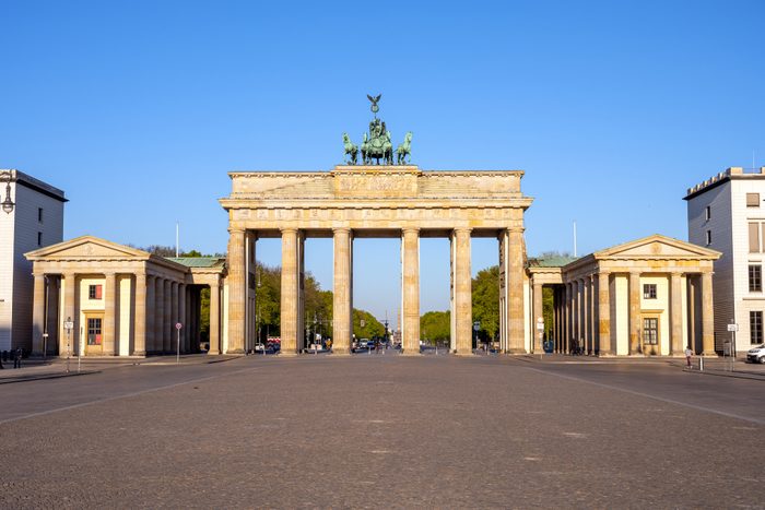 Brandenburg Gate stands tall under a clear blue sky, surrounded by wide cobblestone streets and leafy trees, with no people in immediate view.