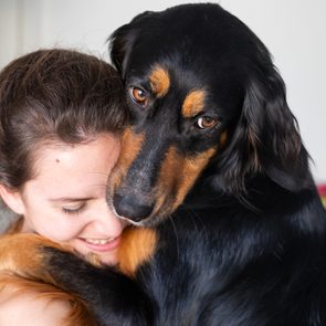 Dog embraces smiling person, its head resting on theirs, in a warmly lit indoor setting with a blurred background.