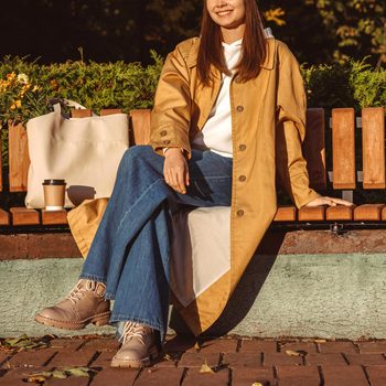A person sits on a wooden bench, wearing a beige coat and jeans, accompanied by a tote bag and coffee cup in a park setting.