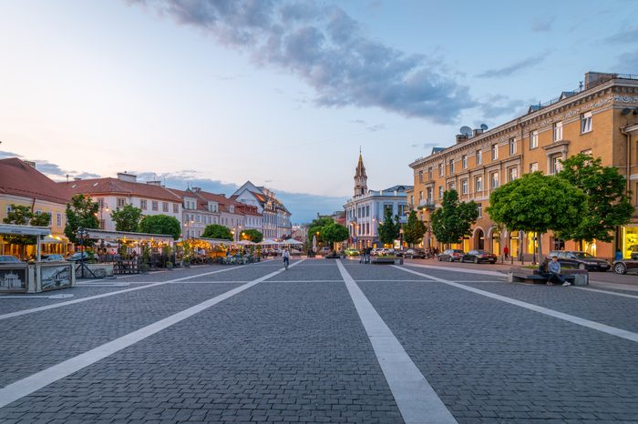 A cobblestone square with people walking and cycling amidst surrounding historical buildings and trees during early evening under a partly cloudy sky.