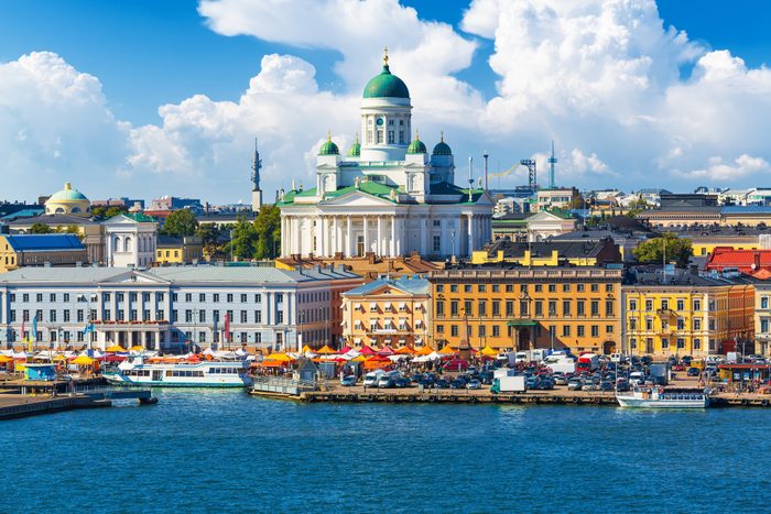 A grand cathedral overlooks a bustling waterfront marketplace with colorful tents, surrounded by historic buildings under a partly cloudy sky.